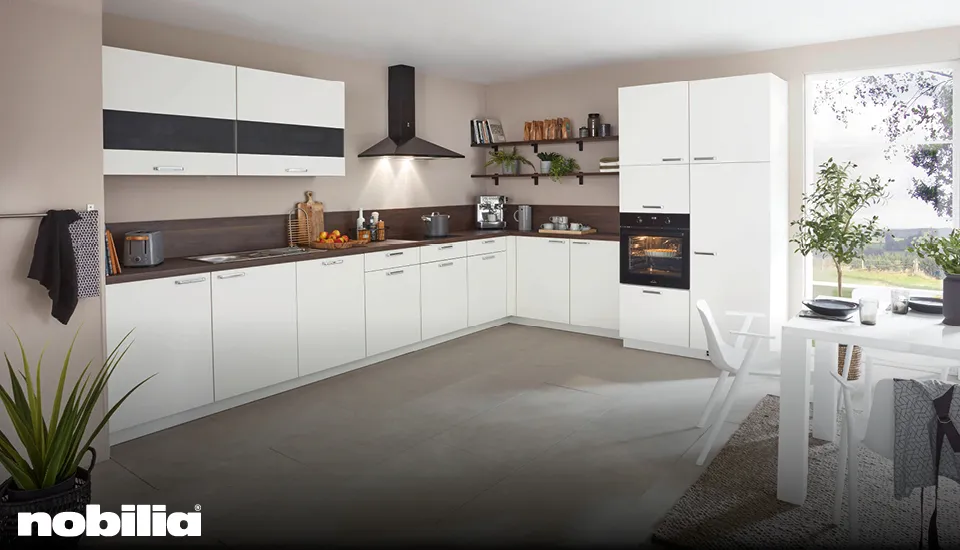 Helle Nobilia Einbauküche in L-Form mit weißen Fronten, dunkler Arbeitsplatte, Hängeschränken mit Glasakzenten und großem Fenster, Essbereich mit weißem Tisch und Pflanzen